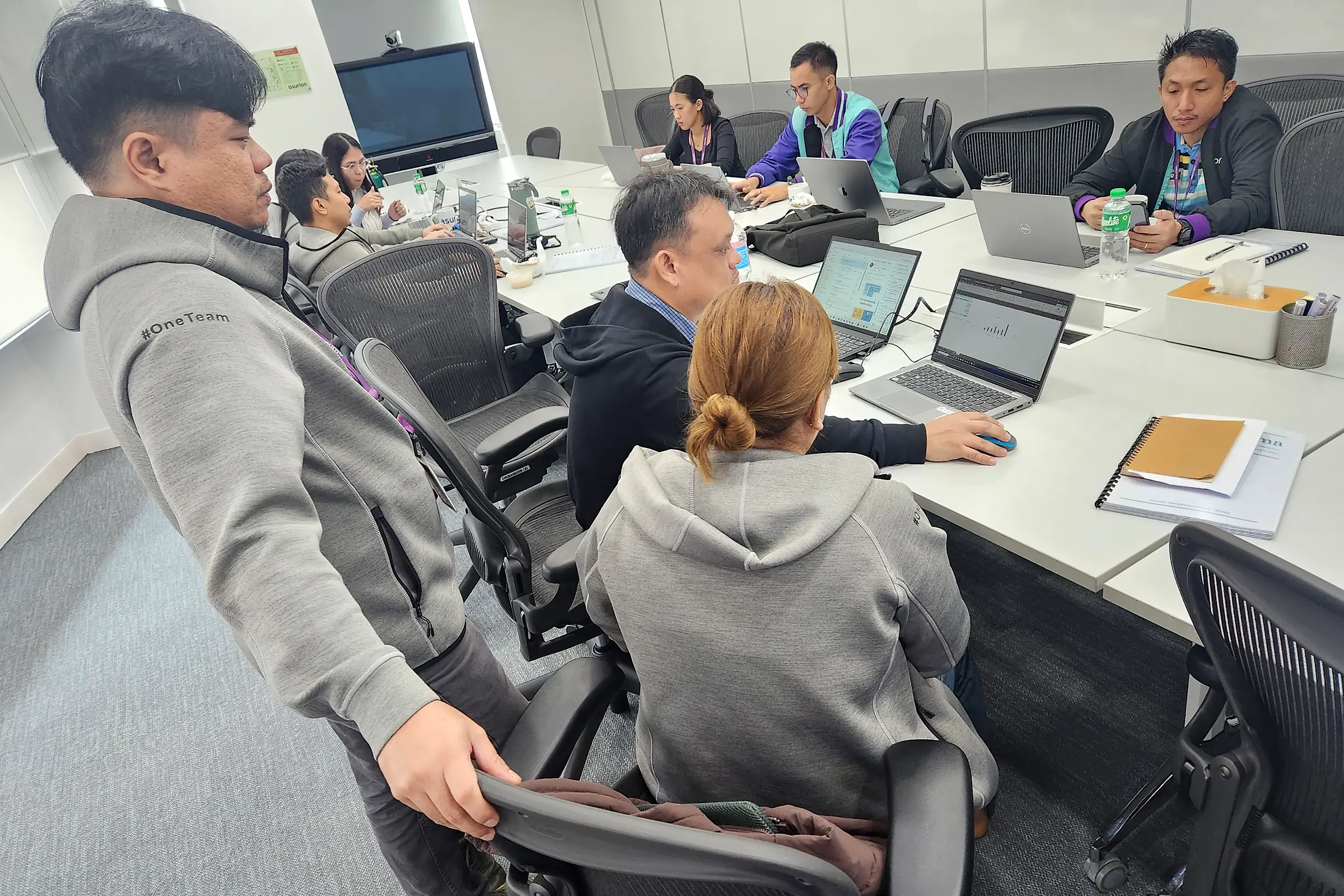 Participants with laptops at a long white table, one man leaning over a colleague's shoulder to look at her screen