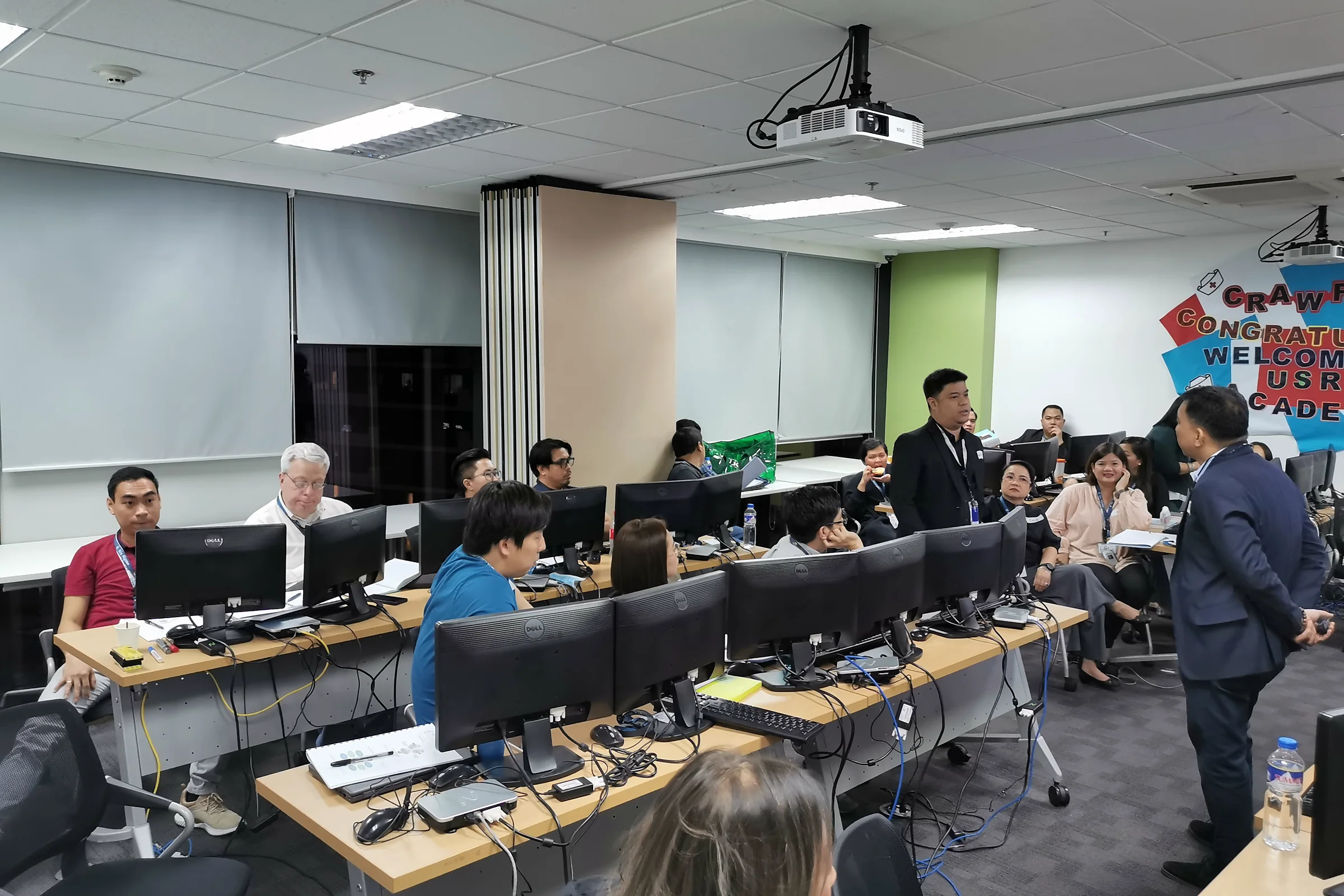 Participants at computer workstations in a classroom while a facilitator stands and talks with them