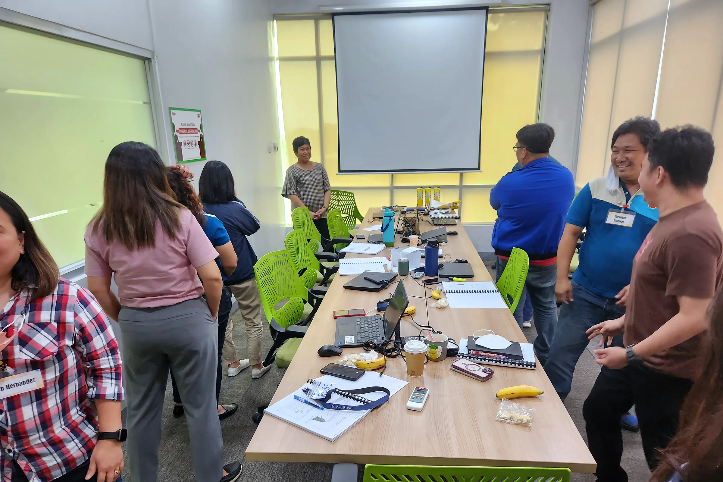 Participants standing around a long table with papers and laptops during a team activity, a projector screen behind them