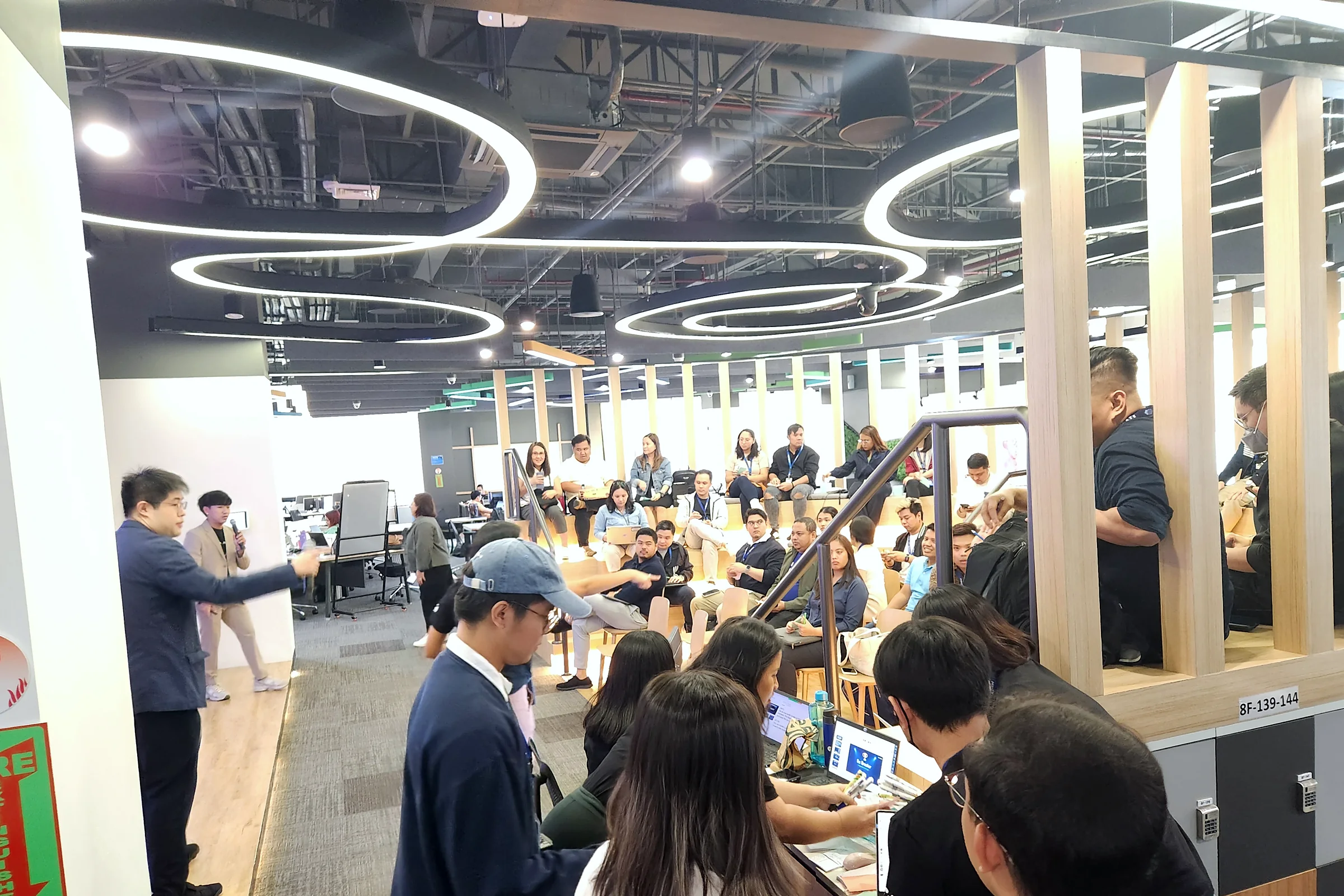 A busy open-plan office event with people seated at tables while a man in a blue shirt speaks at the front