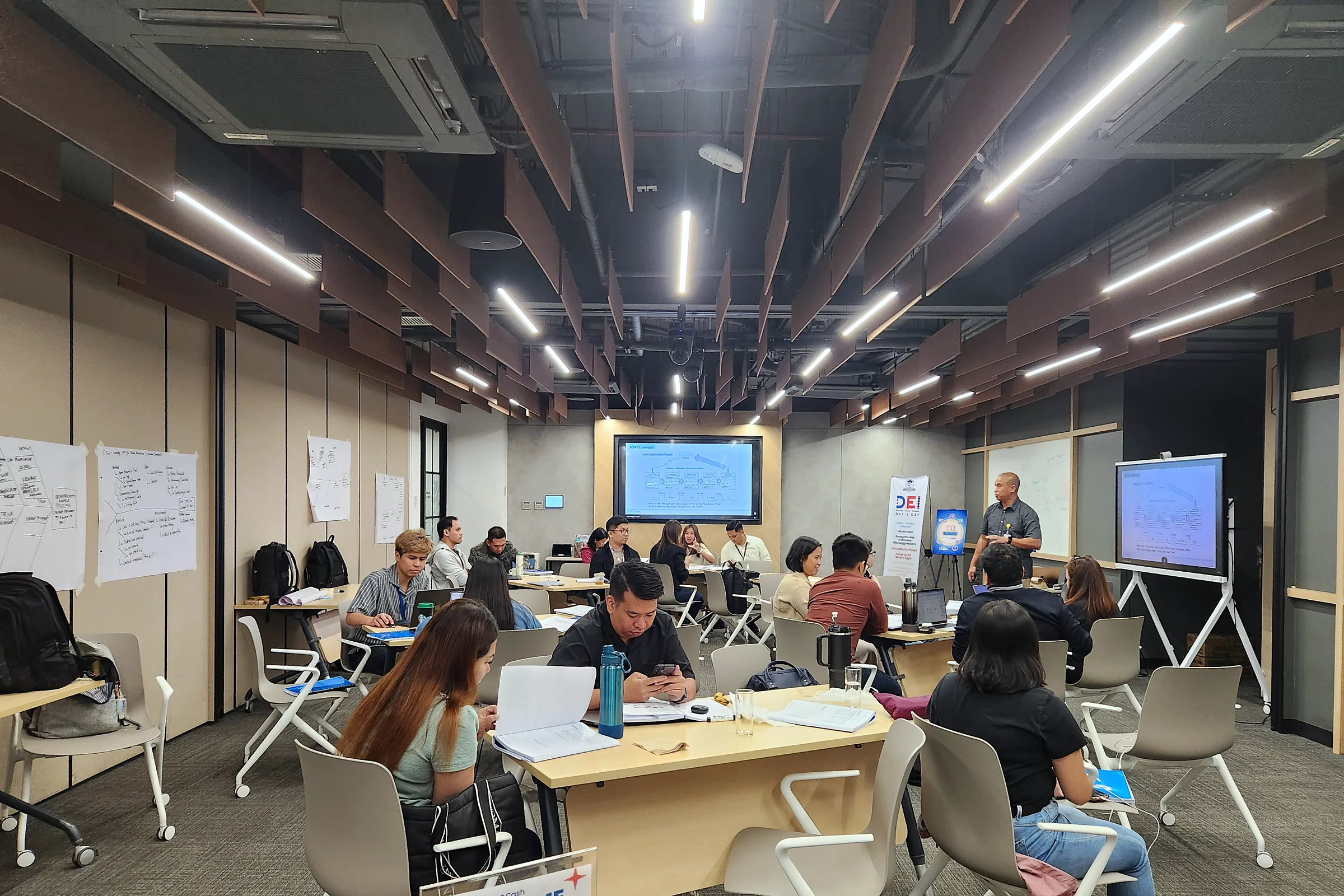 Participants working at tables in a large open office training space, with flip-chart sheets on the walls and screens at the front