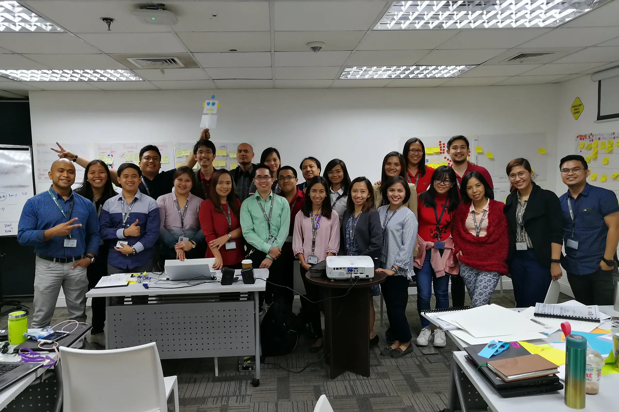 A large group posing together at the back of a training room, some giving thumbs up