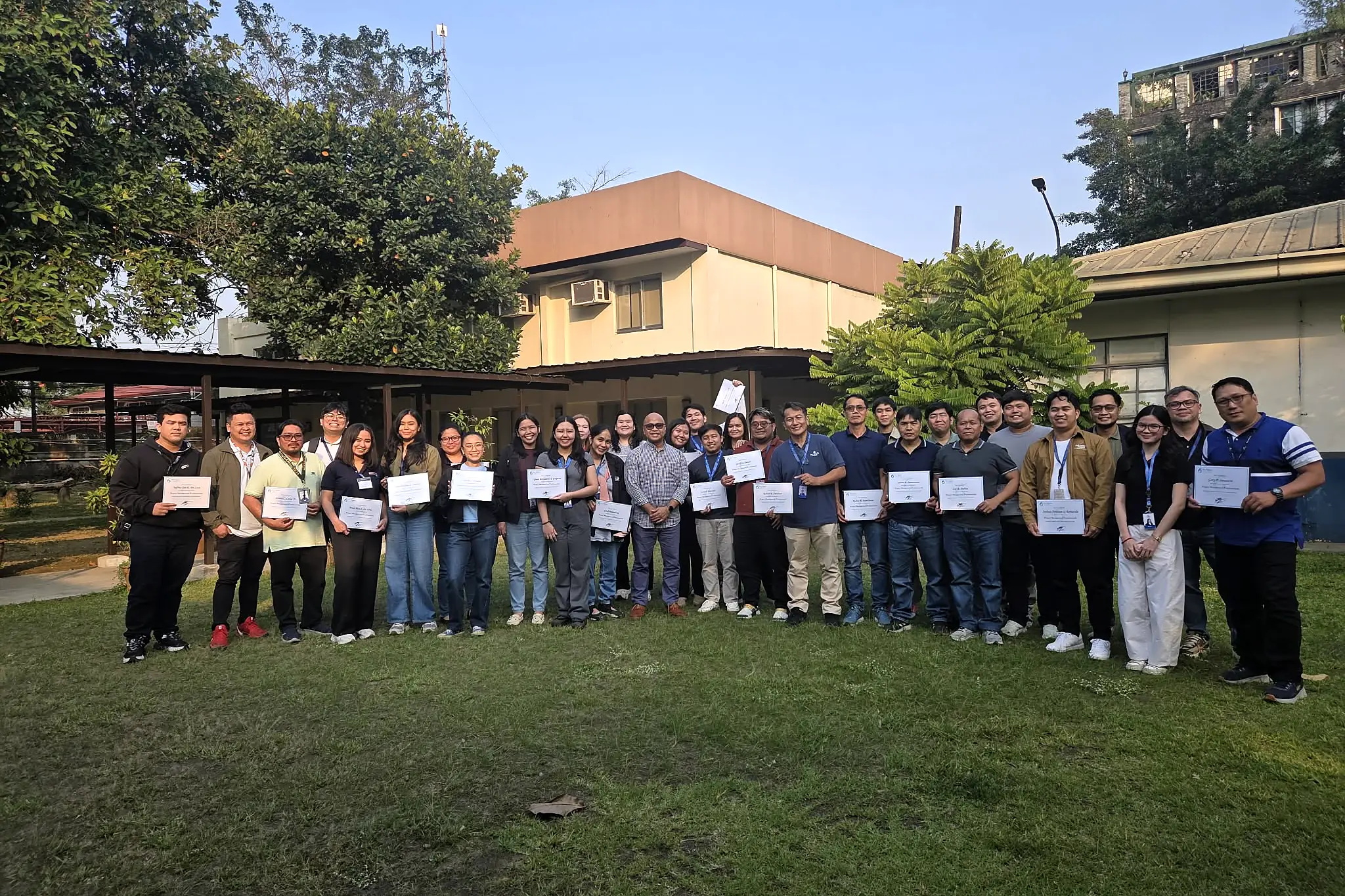 A large class standing outdoors on a lawn in front of a building, each holding up a certificate