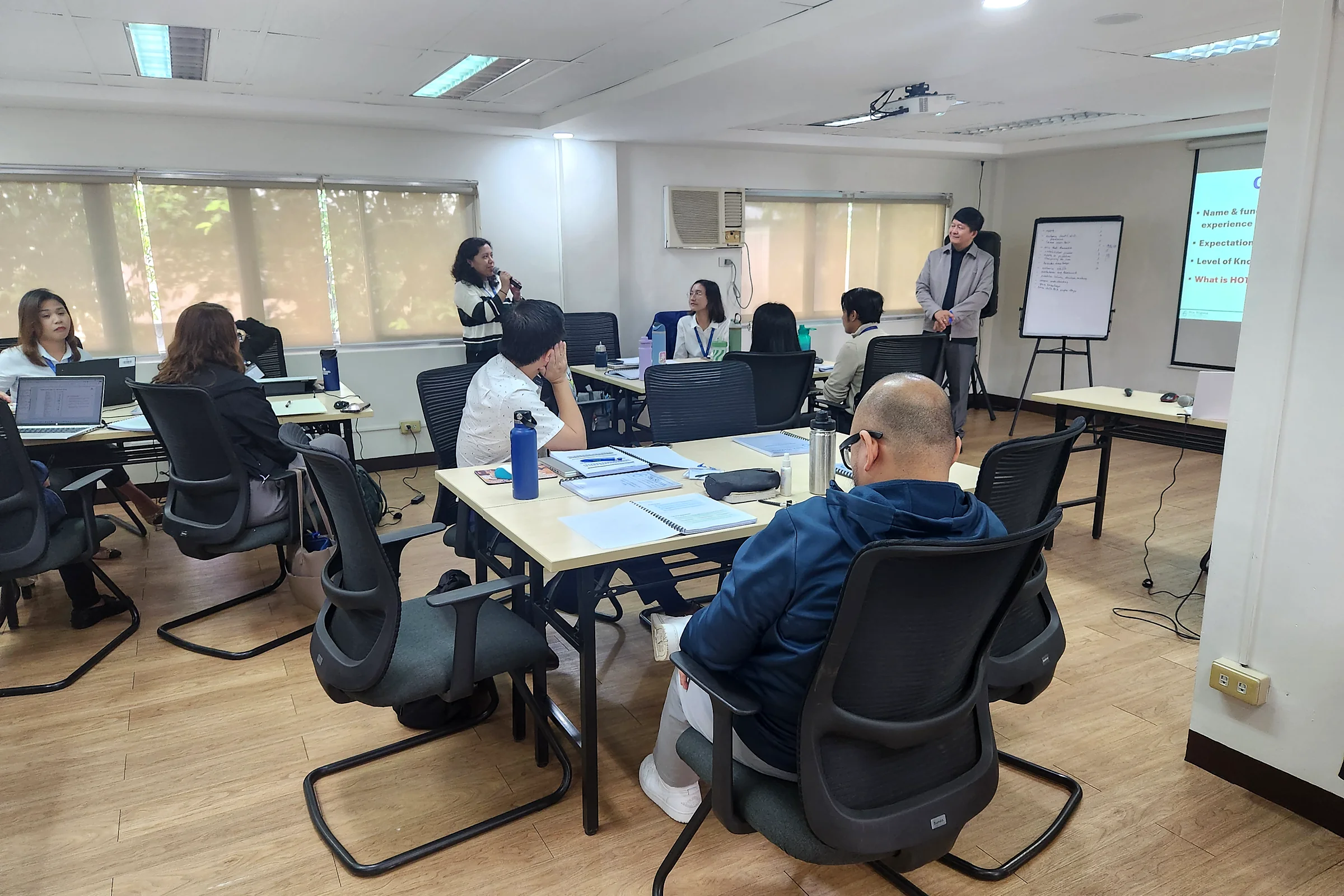 Participants seated at tables in a training room with laptops and flip charts, a facilitator at the front