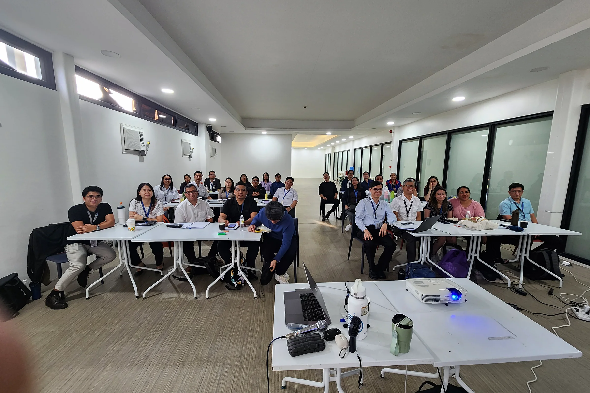 A wide training room with participants seated in rows of white tables facing the front