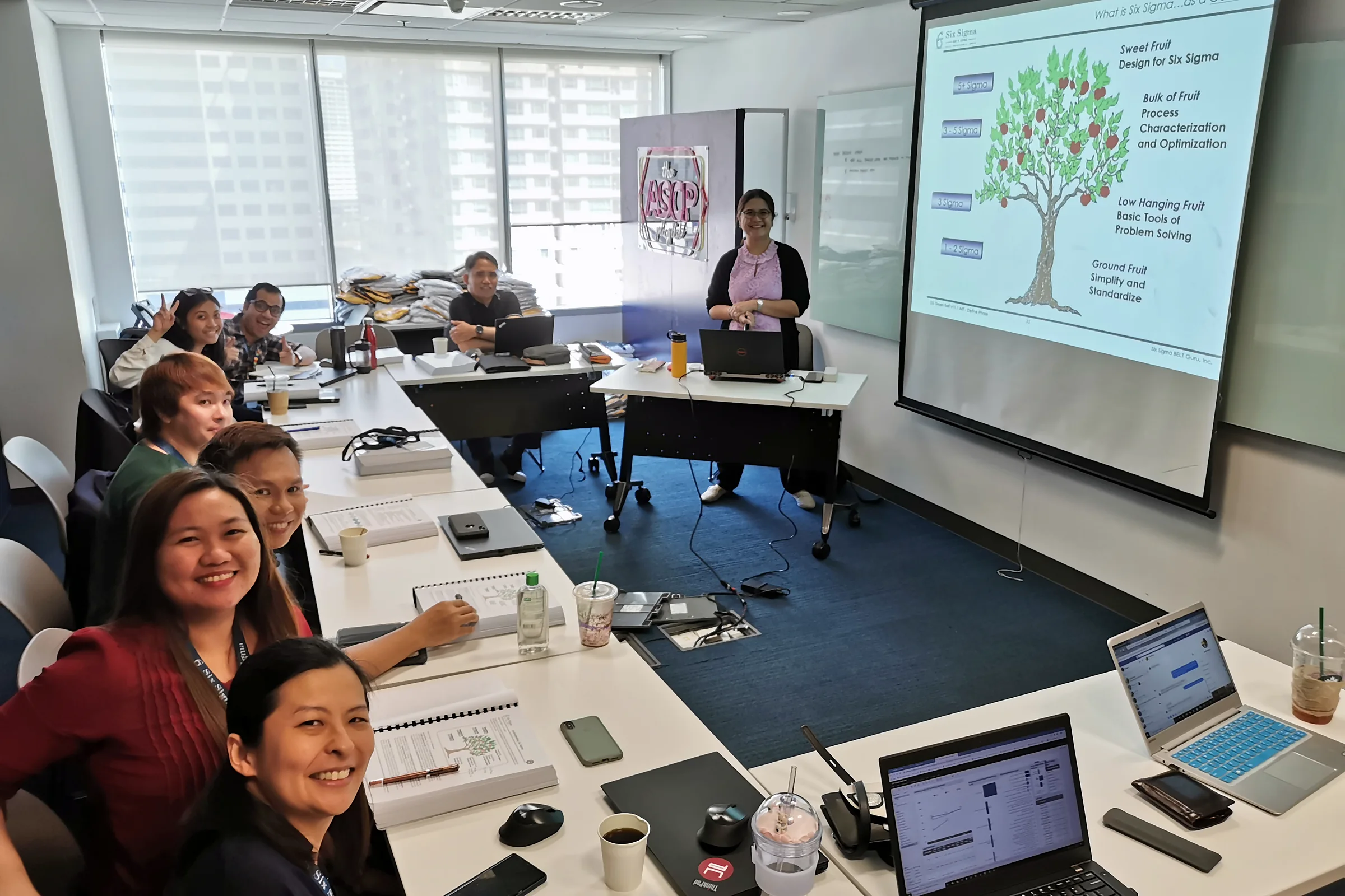 Smiling participants at a U-shaped table with laptops, facing a projected slide of a tree diagram